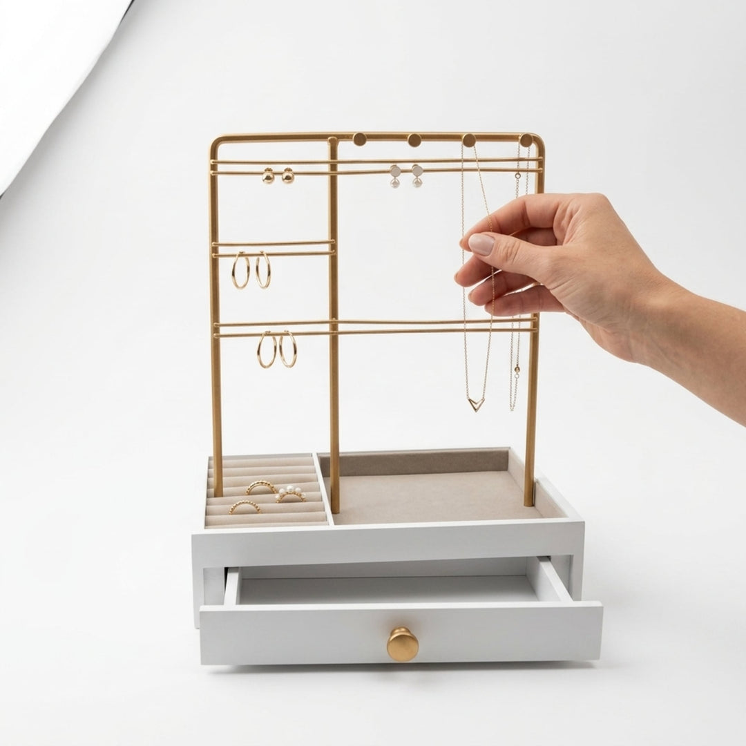 En hånd lægger en halskæde på UNIQ smykkestativ / smykkeskrin i guld med skuffe – Holder til øreringe, ringe og kæder (29 cm) fra UNIQ, der fremviser sine elegante rum og skuffe til praktisk opbevaring af øreringe og ringe på en hvid baggrund.