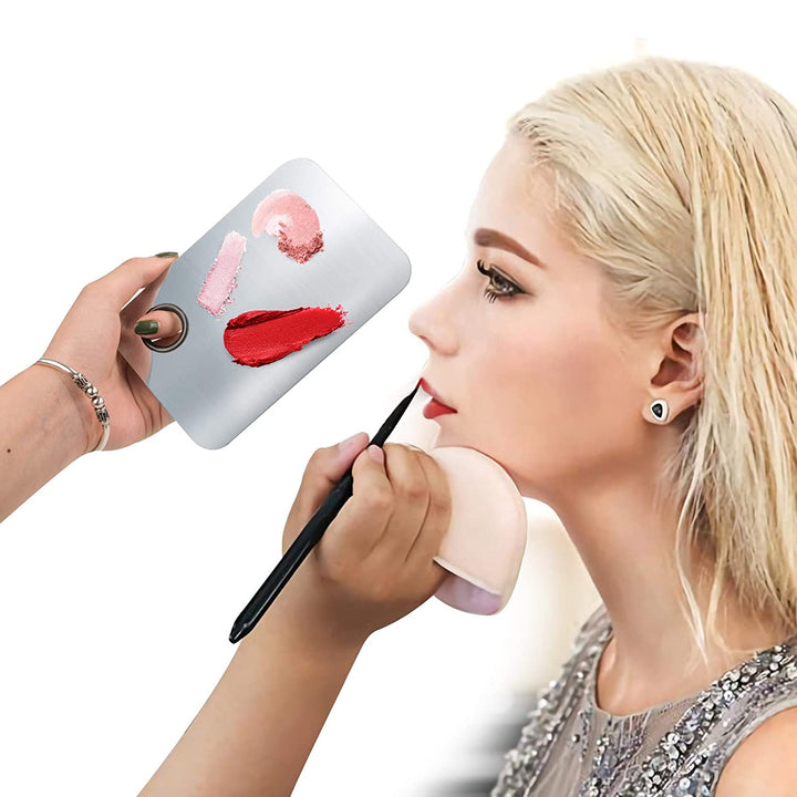 En person påfører læbestift på en blond kvinde med en pensel, mens vedkommende holder Technique PRO Professional Makeup Mixing Palette, der indeholder forskellige nuancer af læbestift. Kvinden har en glitrende top og øreringe på. Denne palet i rustfrit stål fra Technique PRO er ideel til præcis udtoning af læbestift eller foundation under påføring af makeup.