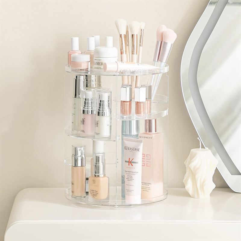 UNIQ 360 graders roterende organizer - Transparent fra UNIQ har plads til hudplejeflasker, makeupbørster, lotion og skønhedsprodukter på en beige bordplade ved siden af et dekorativt lys og et spejl.