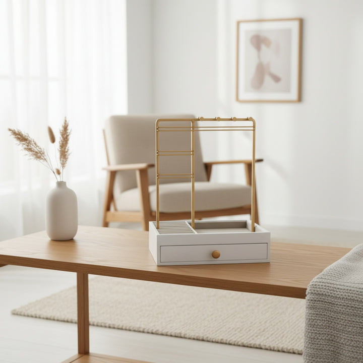 En moderne stue med et sofabord i træ er udstyret med UNIQ Smykkestativ / Smykkeskrin i Guld med Skuffe – Holder til øreringe, ringe og kæder (29 cm) fra UNIQ, der tilbyder stilfuld og elegant smykkeopbevaring med guldskinner og en praktisk skuffe. I baggrunden ses en lys lænestol, indrammet kunst, en vase med tørrede græsser og et stort vindue.