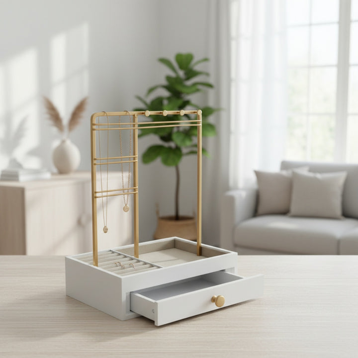 UNIQ Smykkestativ / Smykkeskrin i guld med skuffe fra UNIQ – en elegant holder til øreringe, ringe og kæder (29 cm) – står på et træbord i en lys, moderne stue med store vinduer, en grøn plante og neutrale indretningsfarver.