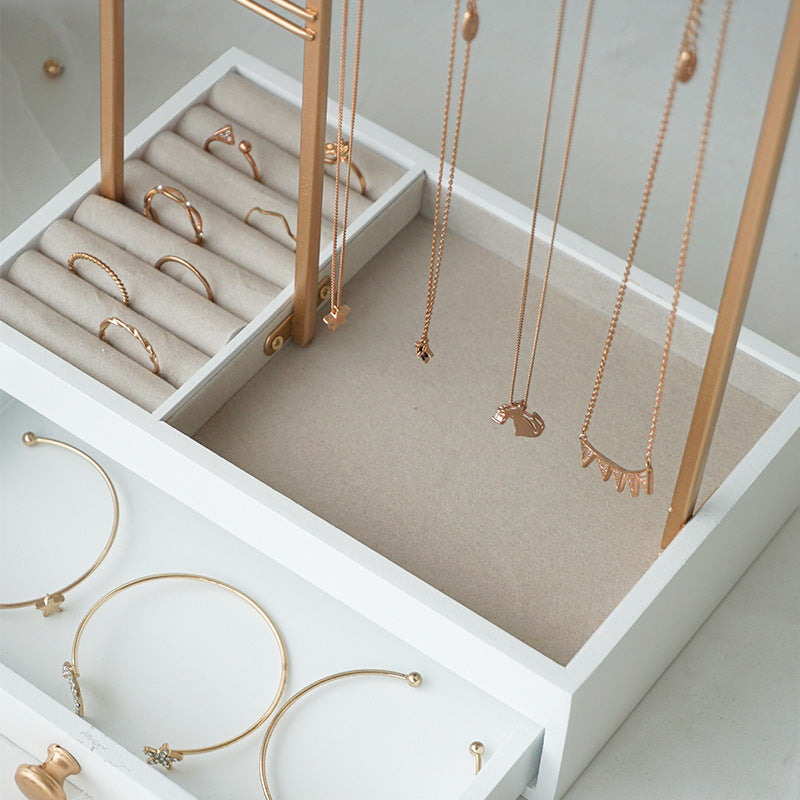 En hvid smykkeskrin fremviser guldringe, armbånd og halskæder, der er anbragt på beige polstrede sektioner, mens halskæderne hænger elegant ned fra UNIQ Smykkestativ / Smykkeskrin i Guld med Skuffe – Holder til øreringe, ringe og halskæder (29 cm) fra UNIQ til stilfuld opbevaring af smykker.
