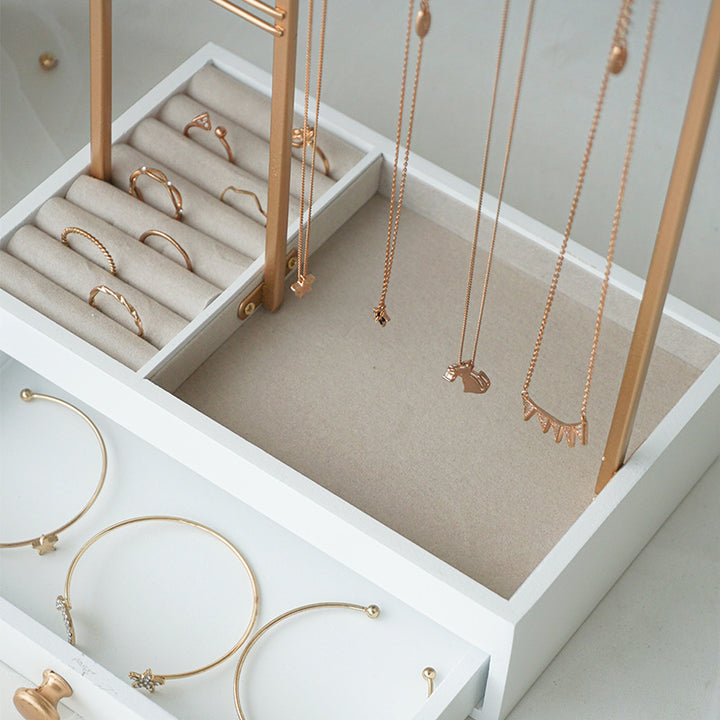 En hvid smykkeskrin fremviser guldringe, armbånd og halskæder, der er anbragt på beige polstrede sektioner, mens halskæderne hænger elegant ned fra UNIQ Smykkestativ / Smykkeskrin i Guld med Skuffe – Holder til øreringe, ringe og halskæder (29 cm) fra UNIQ til stilfuld opbevaring af smykker.